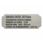 Сребриста гравирана индустриална табелка с надпис „AMADA ENSIS-3015AJc“ и технически спецификации на стационарна лазерна машина