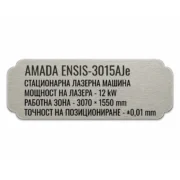 Сребриста гравирана индустриална табелка с надпис „AMADA ENSIS-3015AJc“ и технически спецификации на стационарна лазерна машина